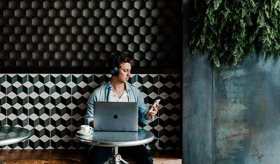 Junger Mann sitzt in einem Café mit Laptop und Smartphone und führt gerade eine digitale Transaktion durch