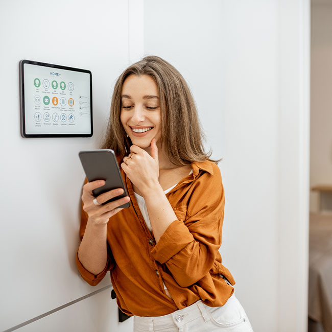 Woman leaning against wall at home accessing smart home platform with her smartphone