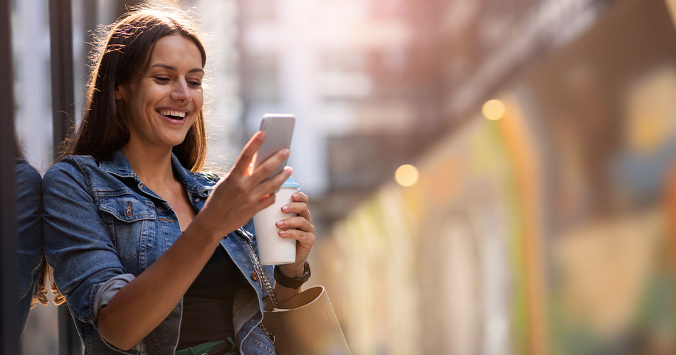 Fröhliche Frau mit Smartphone und Kaffee in der Hand am Straßenrand